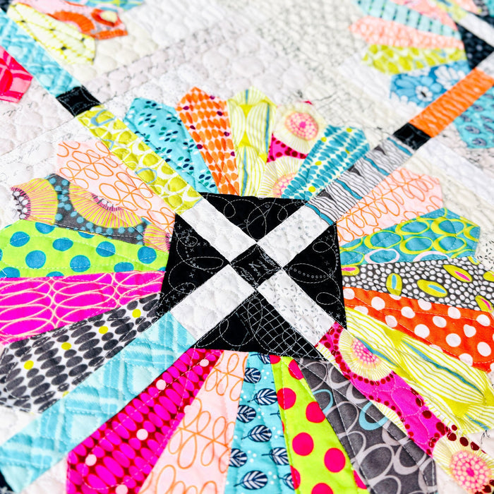 A close-up of a colorful quilted fabric featuring a central black and white checkered square surrounded by brightly patterned radial sections in pink, blue, orange, yellow, green, and gray on a white quilted background.