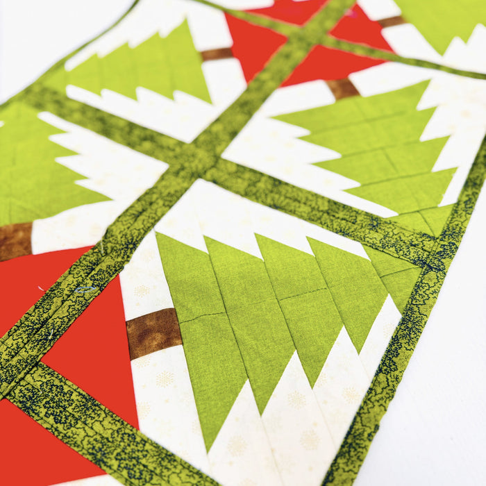 A close-up of a quilted fabric table runner featuring a geometric Christmas tree pattern in green, white, and red, with a decorative olive green patterned border.