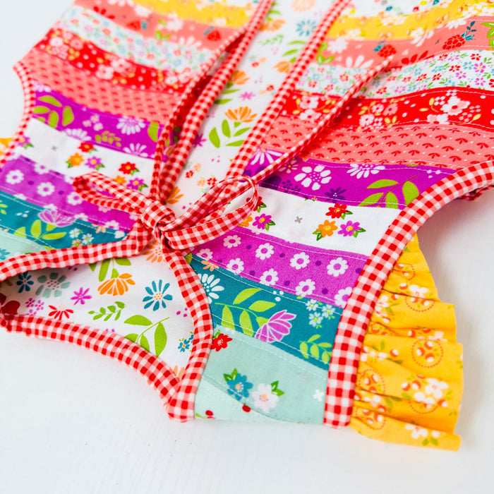 A colorful patchwork apron with a red and white gingham border and tie, featuring various floral and patterned fabric panels in pink, purple, blue, yellow, and red, laid flat on a white background.
