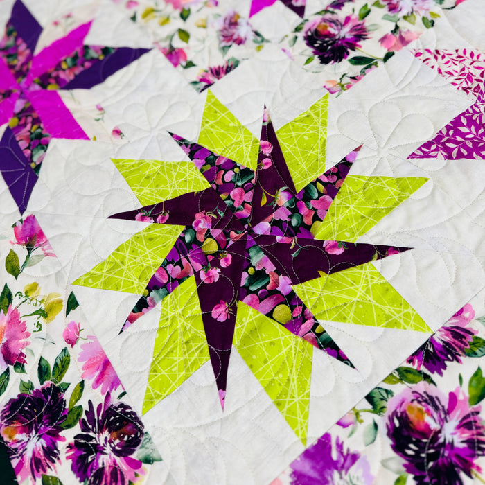 A close-up of a colorful quilted fabric featuring geometric star and floral block patterns in purple, pink, lime green, and white.