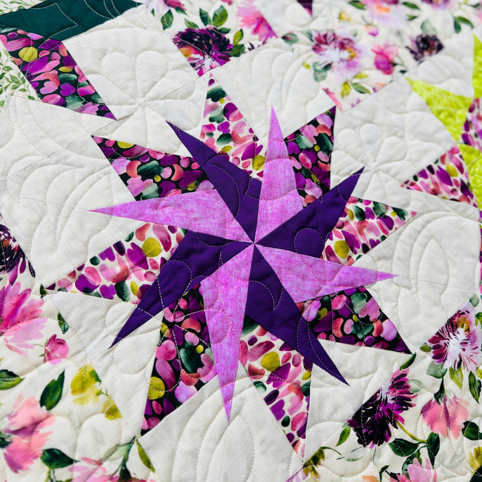 A close-up of a quilted fabric featuring a geometric star pattern in purple and pink, surrounded by white quilted sections and floral prints with green leaves and pink flowers.