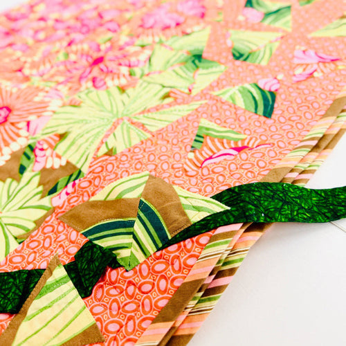 A close-up of a colorful quilted fabric item, likely a quilt or textile panel, featuring a patchwork design with pink floral and green tropical leaf patterns on a coral background, with a dark green ribbon tied around it.