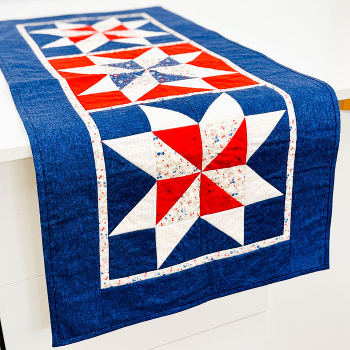 A rectangular quilted table runner with a red, white, and blue patriotic starburst pattern draped over a white table against a white wall.
