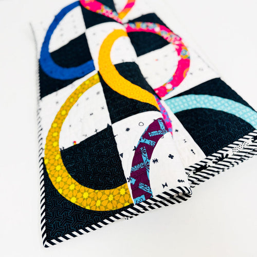 A folded quilted fabric item, likely a small pillow cover or pouch, featuring a modern abstract design with black and white checkered squares and curved stripes in yellow, blue, pink, and light blue, photographed on a clean white background.