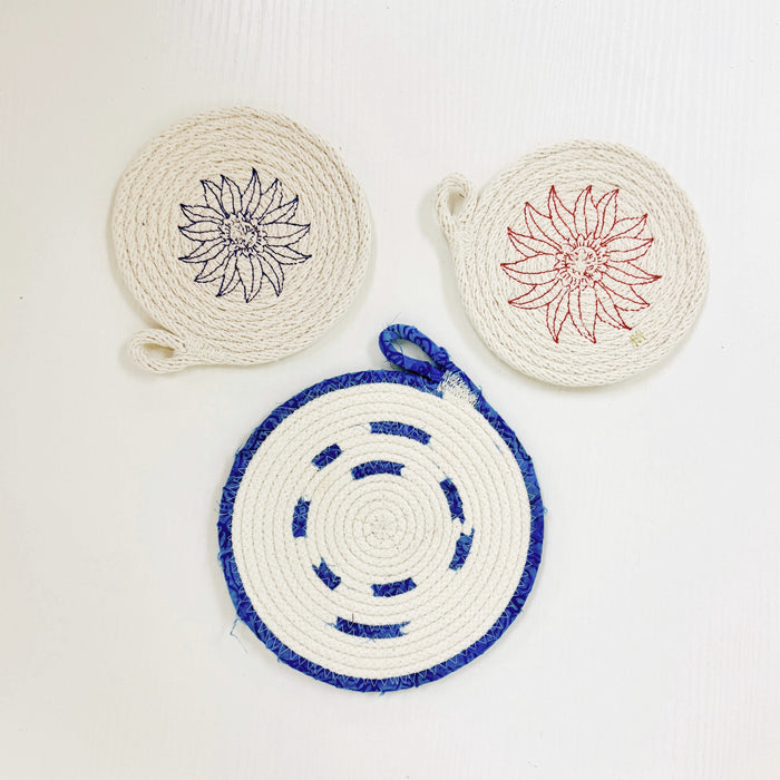 Three round woven rope coasters or trivets on a white background, featuring floral line-art designs in black, red, and blue.
