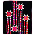 A square quilted textile featuring a black background with four starburst motifs in white and bright pink, red, orange, and purple patterned fabric, connected by vertical striped columns in pink, white, and black.