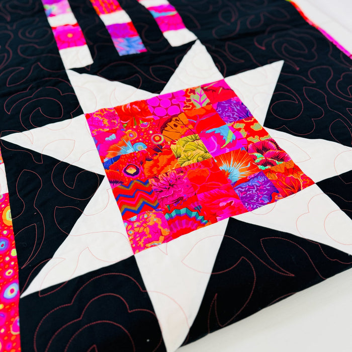 A close-up of a colorful quilted fabric featuring a star pattern with black sections, white points, and a vibrant multicolor patchwork center.